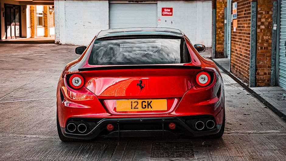 A red Ferrari sports car is parked in an outdoor driveway area, positioned facing away from the camera. The vehicle features a sleek, aerodynamic design with a smooth rear end, circular taillights, an