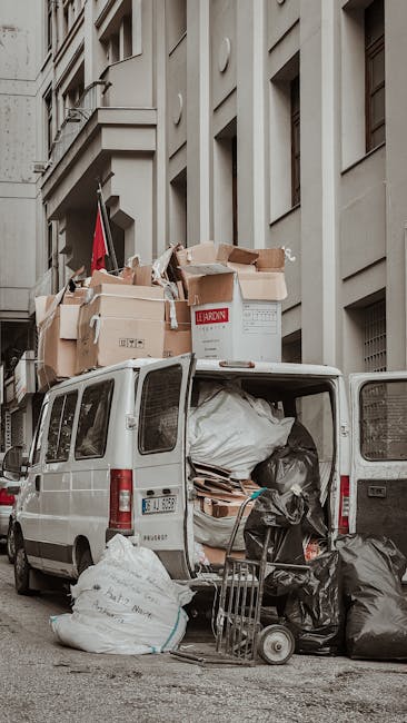 A white commercial van is parked on a city street next to a multi-storey building with large windows and architectural detailing. The back doors of the van are open, revealing filled interior space containing black plastic rubbish bags, cardboard boxes, and packing materials. Several large cardboard boxes are stacked on top of the vehicle, some branded with labels, indicating preparation for a house removal or furniture transport process. A hand truck or trolley is positioned nearby, loaded with additional black rubbish bags and packing supplies. The scene suggests the ongoing loading process associated with home relocation or moving services, with objects placed both inside the van and on the pavement. The environment is urban, with neutral-coloured building facades and diffuse lighting, typical of an active removal or clearance operation managed by Man with Van Newbury Park.