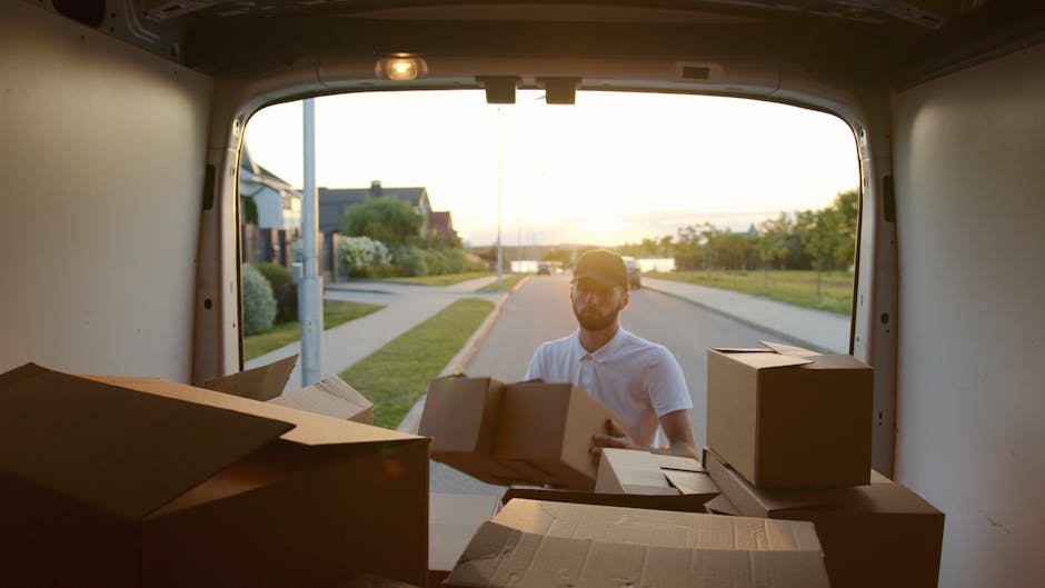 A man wearing a white polo shirt and a cap is seen outside a residential property during daylight, loading medium-sized cardboard boxes into the back of a moving van. The van's rear door is open, revealing several packed boxes, some sealed with packing tape and others open, stacked inside. The van is parked on a suburban street with a sidewalk, greenery, and residential houses visible in the background. The scene is illuminated by natural sunlight, indicating an early evening or late afternoon setting. This image exemplifies a home relocation process managed by Man with Van Newbury Park, showcasing furniture transport and packing tasks as part of a professional removals service.