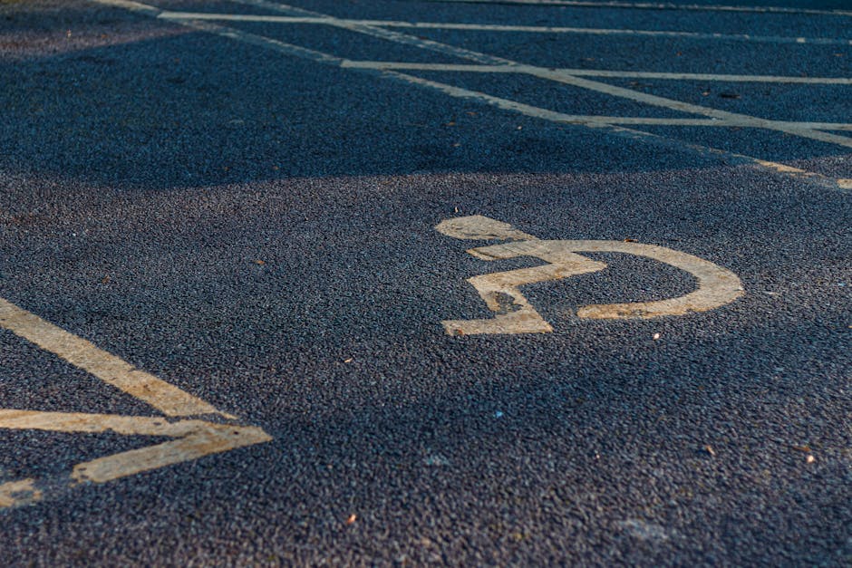 Close-up view of an empty parking space on an asphalt surface with clearly visible white painted lines and a circular symbol with the number '22' inside. The area appears to be part of a designated parking lot adjacent to a residential or commercial property. The surface is textured with small gravel and minor debris scattered around. The background shows additional parking spaces and a small section of the surrounding environment. This setting demonstrates typical parking lot markings relevant for vehicle loading or unloading during home relocation or furniture transport, as managed by firms such as Man with Van Newbury Park, which specialises in removals and moving services in the IG2 postcode area.