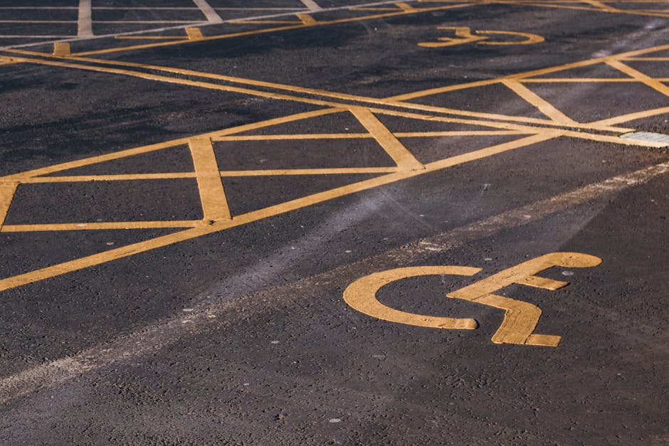 A close-up view of a section of an asphalt parking lot with yellow painted markings for disabled parking, including a wheelchair symbol and grid lines. The surface appears slightly worn with visible cracks and texture. This area is designated for accessible vehicle parking, which may be relevant for home relocation or furniture transport logistics. The parking lot is outdoors and partially lit by natural light. The image is associated with house removals and moving services, highlighting the importance of accessible parking arrangements for efficient loading and unloading during a move, as managed by companies like Man with Van Newbury Park, offering professional removals services within the IG2 postcode area.