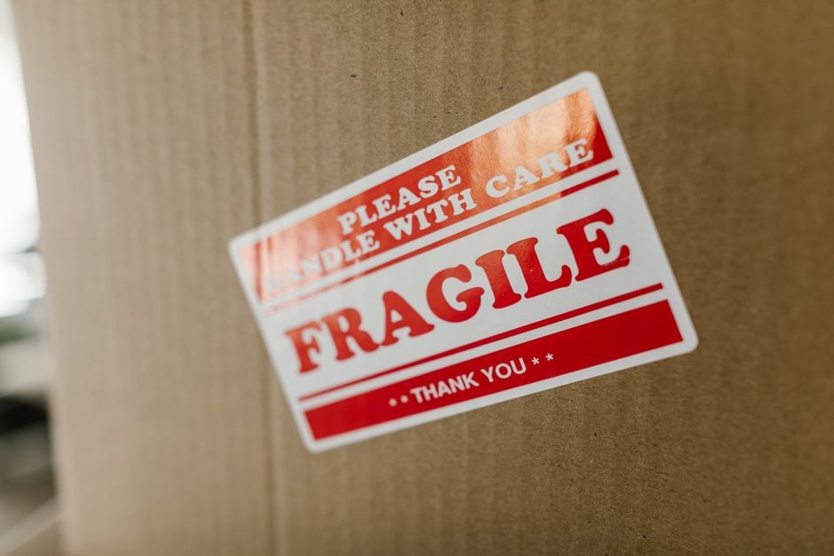 Close-up photograph of a large cardboard moving box inside a property, with a prominent red and white 'Fragile' label affixed to the front. The label includes additional instructions such as 'Please handle with care' and 'Thank you'. In the background, parts of a doorway and interior furnishings are visible, indicating an indoor packing or loading environment. The box is positioned on a flat surface, presumably during a home relocation process managed by Man with Van Newbury Park, highlighting careful handling and transport of delicate items during furniture transport or packing and moving activities.