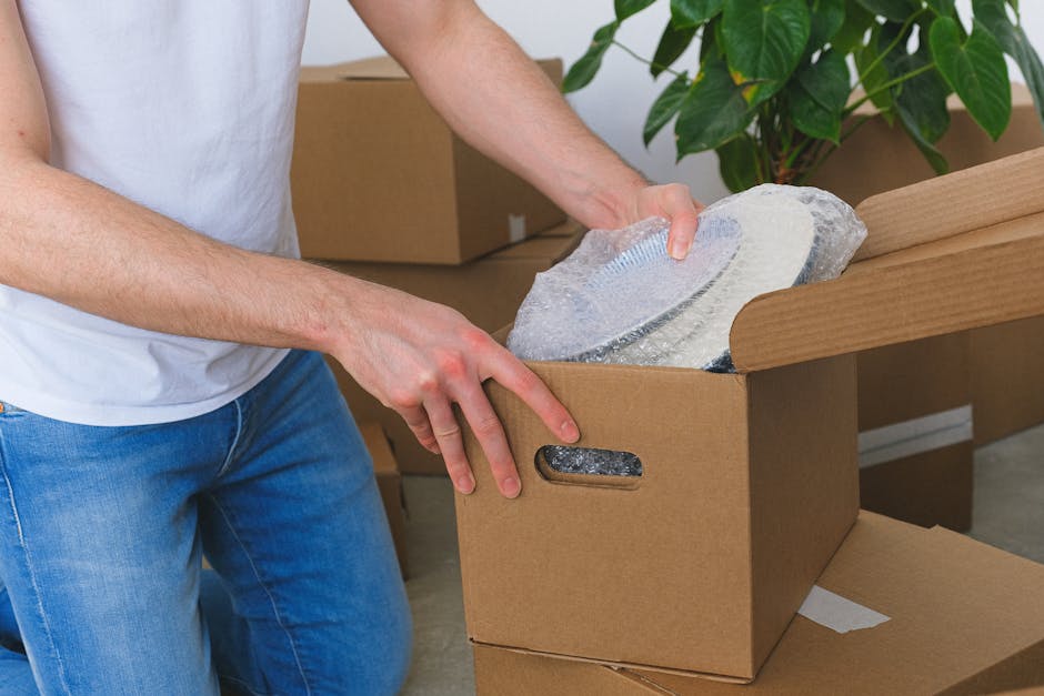A person wearing a white shirt and blue jeans is in the process of packing a fragile item, specifically a glass or ceramic plate, by wrapping it carefully in bubble wrap. They are placing the wrapped item into a cardboard storage box, which is positioned on a flat surface inside a house or storage area. The box has cut-out handles on its sides, and several other boxes, some sealed, are visible in the background. A large green leafy plant is seen near the wall, adding natural decor to the space. The scene depicts careful packing during a home relocation or moving process, highlighting professional packing practices for delicate items, as handled by Man with Van Newbury Park, experts in removals and furniture transport services.