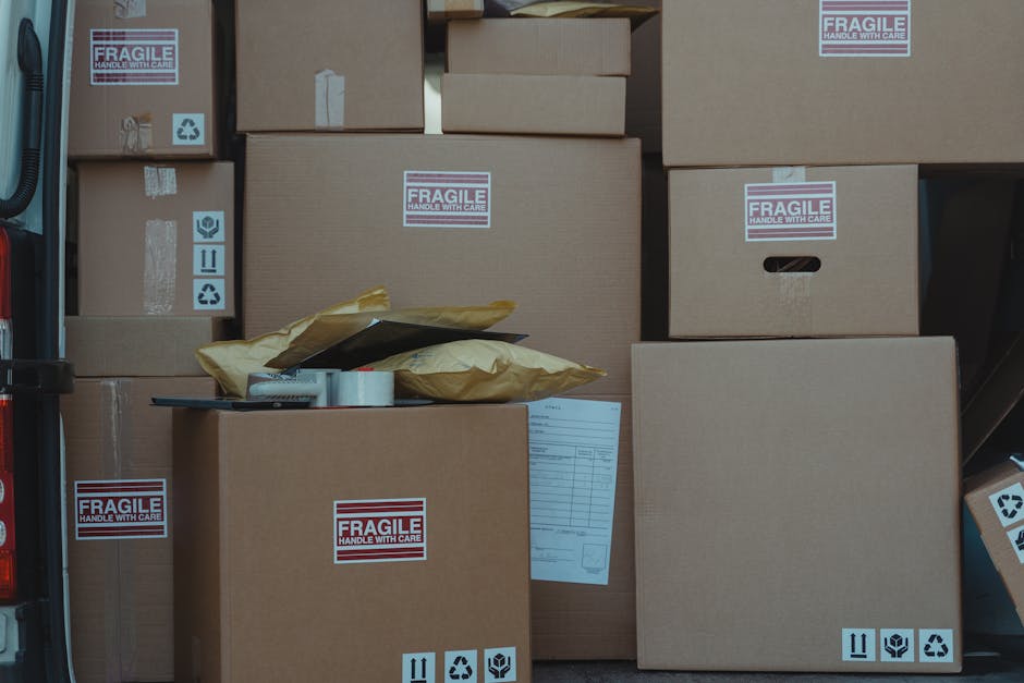 Close-up photograph of a large cardboard moving box inside a property, with a prominent red and white 'Fragile' label affixed to the front. The label includes additional instructions such as 'Please handle with care' and 'Thank you'. In the background, parts of a doorway and interior furnishings are visible, indicating an indoor packing or loading environment. The box is positioned on a flat surface, presumably during a home relocation process managed by Man with Van Newbury Park, highlighting careful handling and transport of delicate items during furniture transport or packing and moving activities.