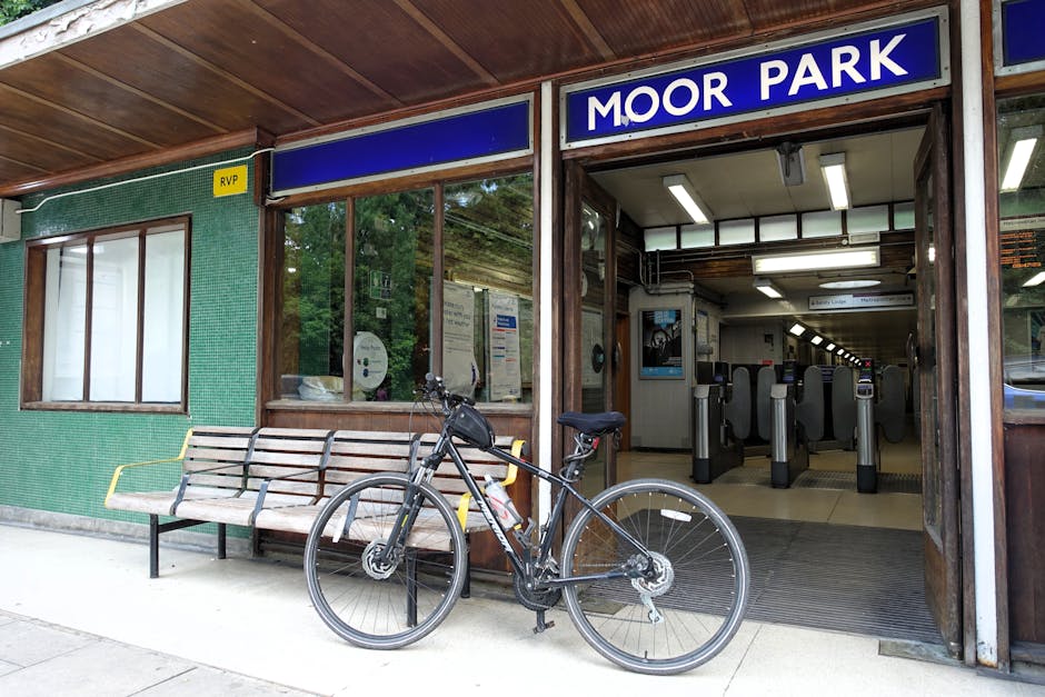 A black bicycle with a water bottle mounted on the frame is parked in front of the entrance to Moor Park underground station, which features an open doorway with automatic ticket barriers visible inside. The station has a wooden overhang ceiling and a green tiled exterior wall with a small, rectangular window and a yellow RVP sign. A wooden bench with a metal frame is positioned alongside the wall, and the scene is illuminated by natural daylight. The interior of the station shows lit fluorescent strips on the ceiling, and posters and notices are posted on the walls inside. This setting relates to house relocations and moving logistics, as Man with Van Newbury Park offers professional removals and packing services to facilitate efficient furniture transport during home or office moves.