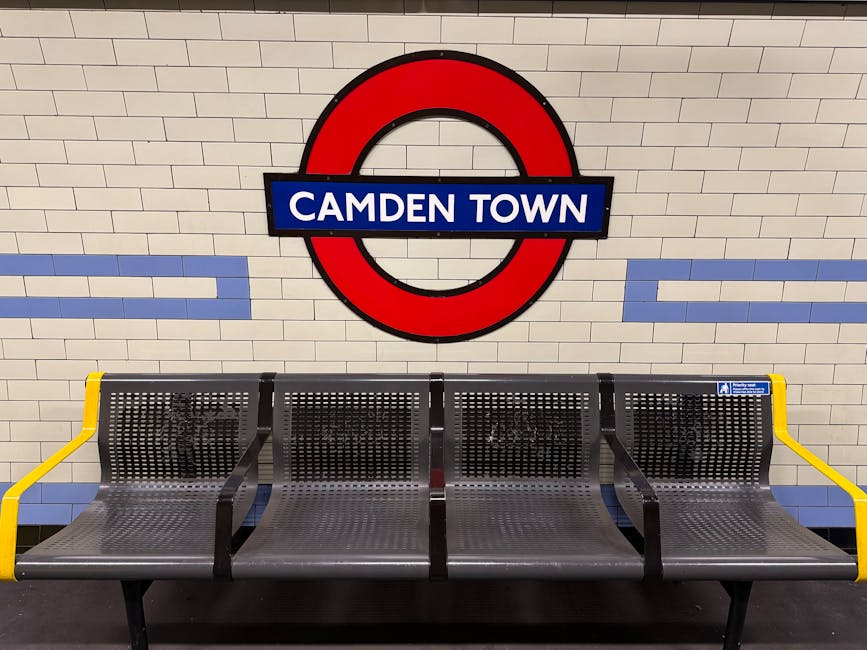 A row of four metal seating benches with perforated surfaces, situated against a tiled wall inside Camden Town underground station. The benches have yellow armrests on both ends and are positioned on a dark platform floor. Above the benches, a large circular London Underground sign features a red circle with a blue bar across the middle displaying the words 'Camden Town' in white text. The station lighting illuminates the area evenly, providing a clear view of the seating and signage. This setting depicts an interior typical of the station environment, relevant to travel and transportation in the context of house removals or home relocation services offered by Man with Van Newbury Park, emphasizing the station's infrastructure for commuters.