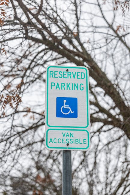 A reserved parking sign indicating it is designated for disabled access and van parking, mounted on a metal pole against a background of leafless tree branches and an overcast sky. The sign features a blue and white wheelchair symbol with the words 'RESERVED PARKING' at the top and 'VAN ACCESSIBLE' at the bottom. This type of signage is commonly used in residential areas, such as Newbury Park, to reserve parking spaces for moving vans or accessible vehicles during home relocation or furniture transport activities. The image relates to the process of arranging parking permits for Newbury Park movers, such as those provided by Man with Van Newbury Park, to facilitate smooth home removals and packing and moving services in the area.
