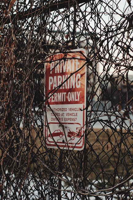 A red and white 'Private Parking Permit Only' sign attached to a metal fence overgrown with dry, tangled brown vines. The sign indicates that parking is restricted to authorized vehicles only and warns that unauthorized vehicles parked at the owner's expense may be towed. The background shows a partly cloudy sky with soft lighting, suggesting an outdoor setting near a property or loading area where house removals or furniture transport activities may take place. This scene highlights the importance of parking permits during home relocation or moving services, with Man with Van Newbury Park ensuring compliance with parking regulations when facilitating moves or furniture transport.