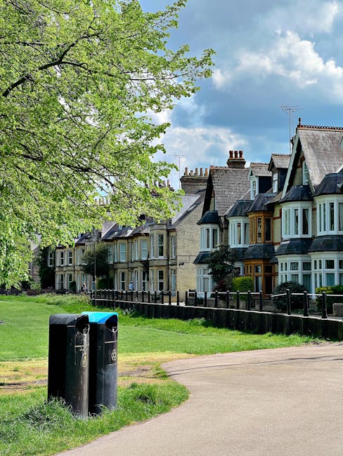 A row of multi-story Victorian-style houses with bay windows and pitched roofs located along a residential street, with a large leafy green tree on the left side partially overhanging the scene. In the foreground, two black wheelie bins are positioned on a paved sidewalk next to a grassy verge. The street appears calm and well-maintained, with the houses set behind a low black fence. The sky above is partly cloudy with patches of blue, suggesting daytime. This setting illustrates a typical suburban neighbourhood where home relocation and furniture transport services, such as those provided by Man with Van Newbury Park, might be utilised during house removals or packing and moving processes, often requiring careful handling of household belongings within tight urban spaces.