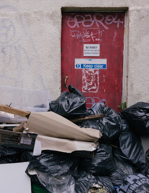 A pile of black garbage bags and scattered discarded packaging materials, including cardboard boxes and plastic wrapping, stacked against a weathered red metal door covered with graffiti and stickers, located next to a textured beige wall. The door has signs indicating no smoking and keeping clear, with one sign referencing bulky waste rules. The trash is situated on a pavement area outside a property, with some debris overlapping the base of the door frame. Visible in the scene are items associated with house removals or clearing, reflecting waste collection and disposal processes prior to moving or clearance, with the backdrop of urban environment and typical external property features. Man with Van Newbury Park would handle such waste as part of their house removal or packing and transport services, ensuring proper disposal and site clearance as part of their moving logistics.