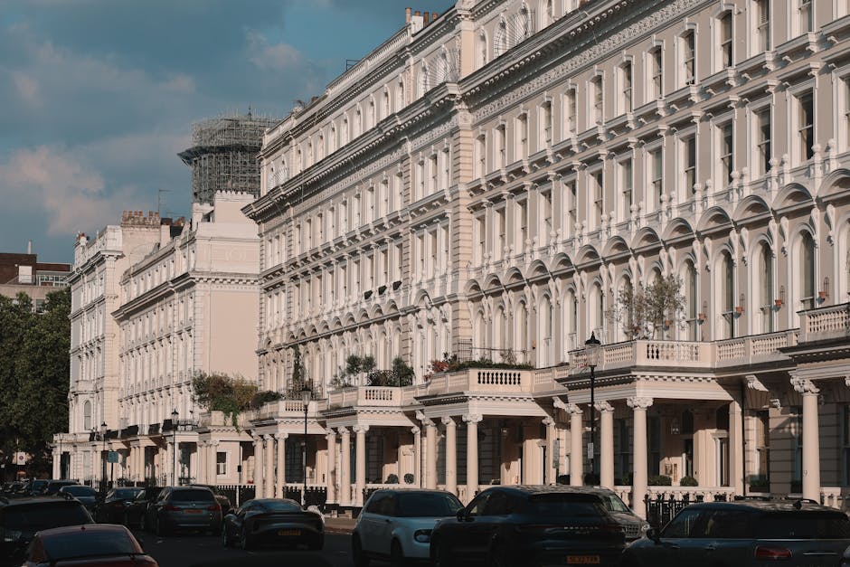 A row of Victorian terraced houses with white facades and ornate architectural details, situated on a busy urban street. In the foreground, multiple parked cars line the pavement, with some partially 