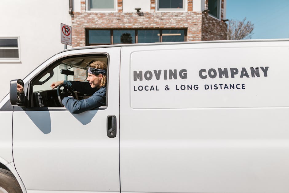 A white moving van with the branding 'MOVING COMPANY LOCAL & LONG DISTANCE' parked on a residential street, with a professional mover inside the cab adjusting controls. The vehicle is positioned next to a house with a brick facade and large windows, indicating a home relocation setting. The van's side door is closed, and nearby on the pavement are cardboard boxes, furniture blankets, and packing materials prepared for the loading process. Visible behind the van are a pavement area and a 'No Parking' sign. Bright daylight illuminates the scene, emphasizing the logistics involved in packing, loading, and transporting belongings for a house move, as handled by a company that offers professional removals services.